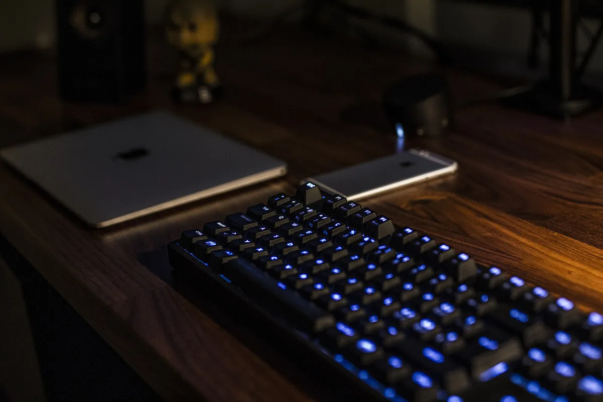 Klawiatura z niebieskim podświetleniem na ciemnym drewnianym biurku, obok otwarty laptop